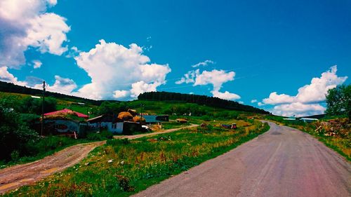 Road amidst field and buildings against sky