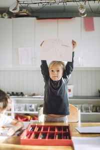 Portrait of girl showing paper with drawing while standing near bench in classroom