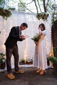 Full length of newlywed couple laughing and standing near wedding arch decorated with flowers and light bulb garlands while reading vows