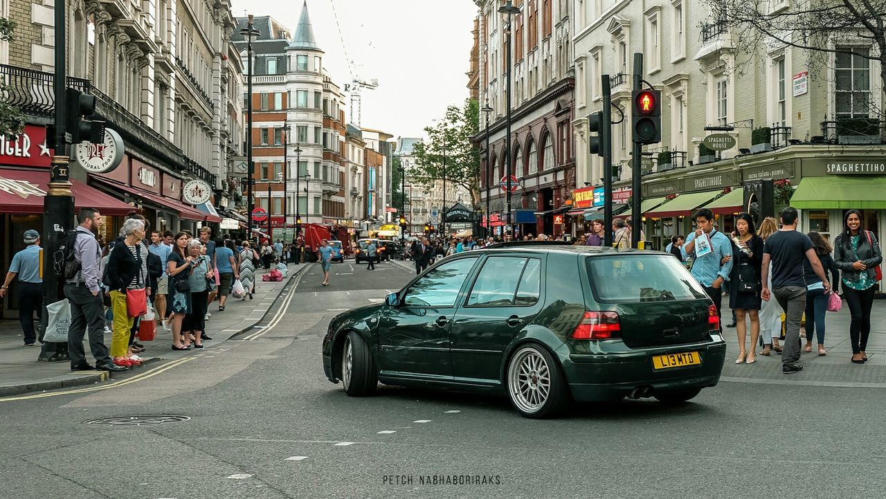 transportation, street, land vehicle, car, building exterior, mode of transport, city, architecture, large group of people, men, built structure, city life, person, road, city street, traffic, walking, on the move, lifestyles