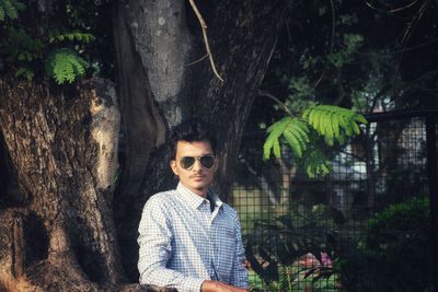 Portrait of young man in sunglasses standing against tree trunk