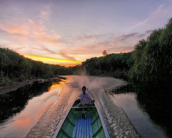 Rear view of man sitting on lake against sky during sunset