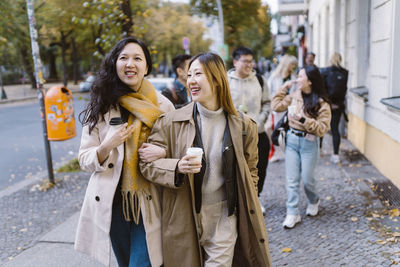 Smiling female friends walking with arm in arm while walking on sidewalk