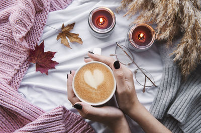 Female hands holding a cup of hot coffee, autumn flat, with yellow leaves, cozy knitwear, candles