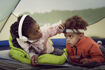 Sister adjusting headlamp for brother while lying with digital tablet in tent