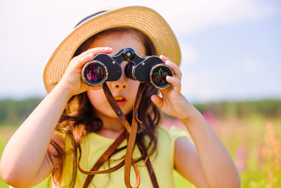Cute girl, in a straw hat, stands in nature, against the blue sky, looks through binoculars 