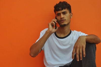 A indian guy making phone call with looking sideways, sitting against orange wall with copy space 