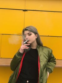 Portrait of young woman smoking cigarette against yellow wall