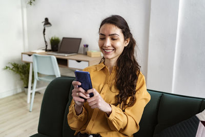 Young woman smiling while using mobile phone sitting on sofa at home