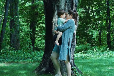 Side view of pregnant woman carrying son while standing by tree trunk in forest