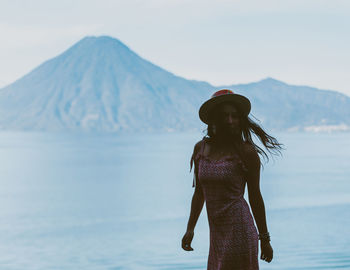 Woman wearing hat standing against sea and mountain
