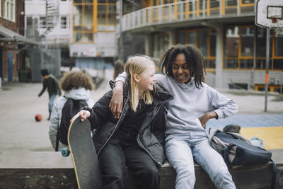 Smiling girl holding skateboard while sitting with friend on retaining wall at school campus