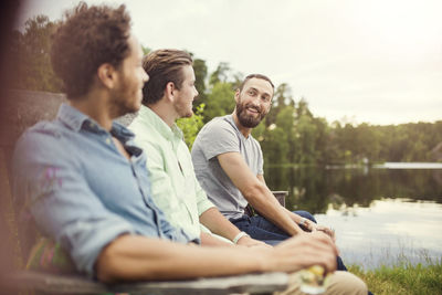 Happy male friends talking while sitting on wooden bench at lakeshore