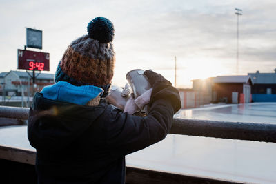 Boy holding ice skates looking out to an empty ice rink at sunset