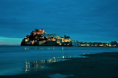 Illuminated buildings by sea against sky at dusk