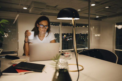 Mature businesswoman holding coffee cup while working late at illuminated creative workplace
