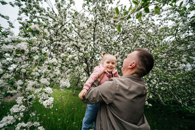 Dad holds a joyful baby daughter in his arms in a garden with a blooming apple tree