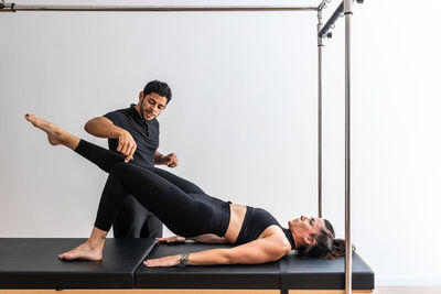 Slim female in sportswear doing exercises on cadillac reformer with help of professional trainer during pilates workout