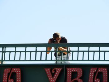 Man photographing while standing by railing against clear sky