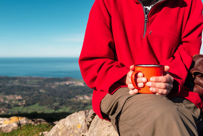 Unrecognizable crop explorer sitting on rock in highlands with metal mug of hot drink while relaxing during hiking in el mazuco