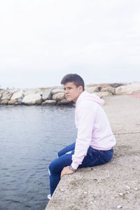 Side view of young man sitting on retaining wall by sea against clear sky