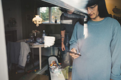 Woman using smart phone with man and baby in background seen through glass window