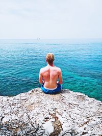 Rear view of shirtless man looking at sea while sitting on cliff against sky