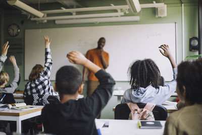 Rear view of multiracial students raising hands in classroom during lecture