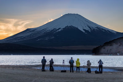 People standing on riverbank against snowcapped mountains during sunset