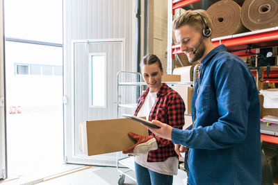 Smiling man showing digital tablet to female coworker holding box in distribution warehouse