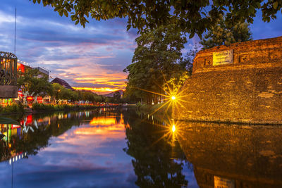 Illuminated buildings by lake against sky at sunset