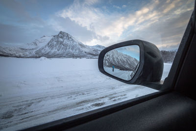 Person on snow covered field reflecting over side-view mirror of car