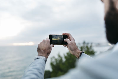 Cropped hands of man photographing sea from mobile phone