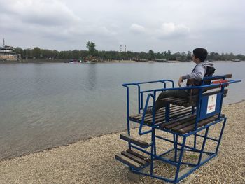 Boy sitting on riverbank against sky