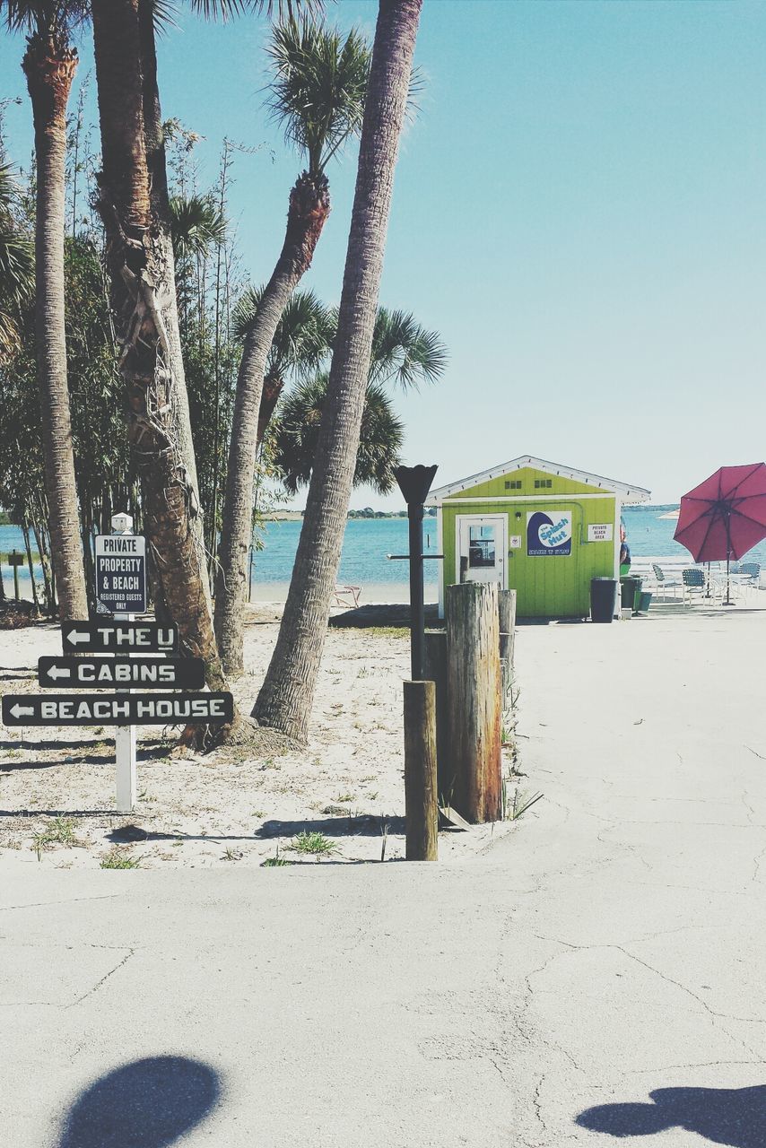 clear sky, text, beach, western script, sand, blue, tree, communication, built structure, sea, sign, day, architecture, information sign, shore, sunlight, tranquility, nature, outdoors, no people