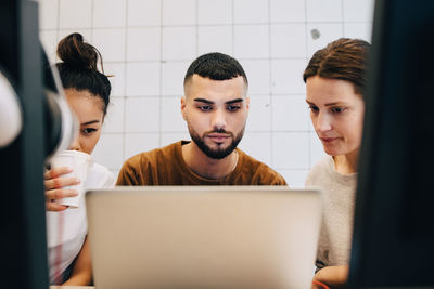 Confident young multi-ethnic colleagues looking at laptop against wall in office