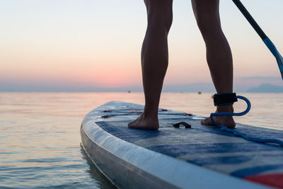 Back view of crop anonymous woman standing on surfboard and rowing with paddle during training on background of amazing sundown sky