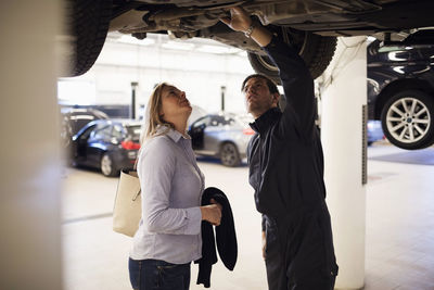 Male expert with client standing under car at repair shop