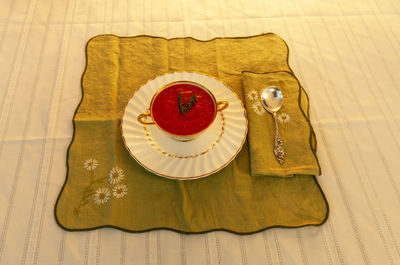 Tomato soup in a white and gold bowl on an embroidered green placemat.