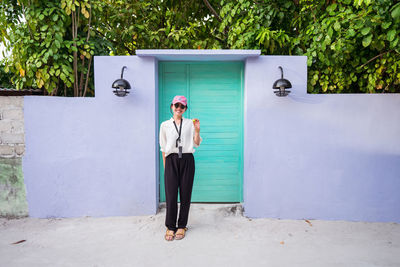 Full body happy young asian female tourist standing near colorful door and waving hand while spending summer holidays on maldives islands
