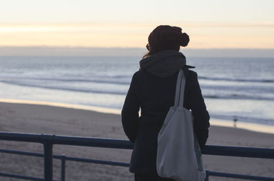 Rear view of woman standing by railing while looking at beach during sunset