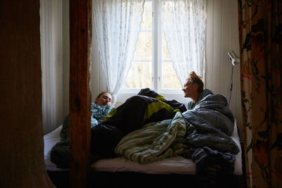 Male and female friends talking while relaxing on bed against window at home