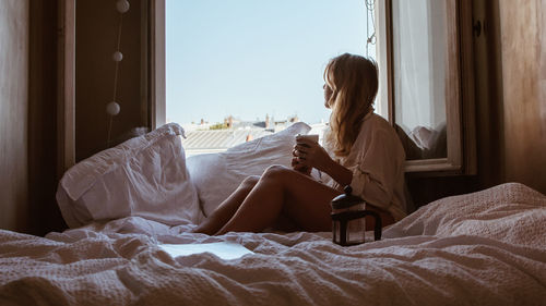 Side view of woman looking through window while sitting on bed at home