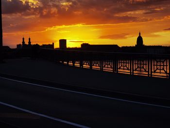 Silhouette buildings by road against sky during sunset