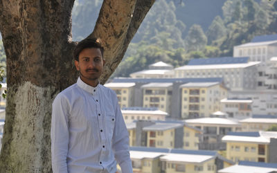 A college student looking at camera standing against tree with college campus in background 