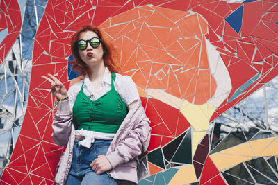 Portrait of redhead young woman wearing sunglasses while gesturing peace sign against mosaic wall