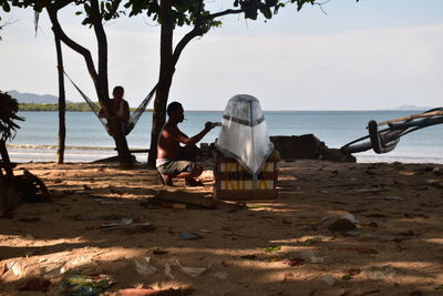 Man painting boat with person sitting on beach by sea against sky
