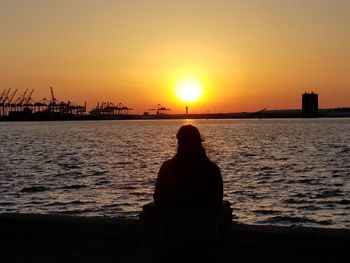 Rear view of silhouette woman sitting by sea against sky during sunset