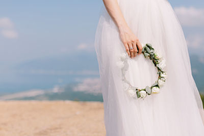 Midsection of bride holding wreath against sky