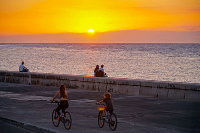 People riding bicycle by sea against sky during sunset
